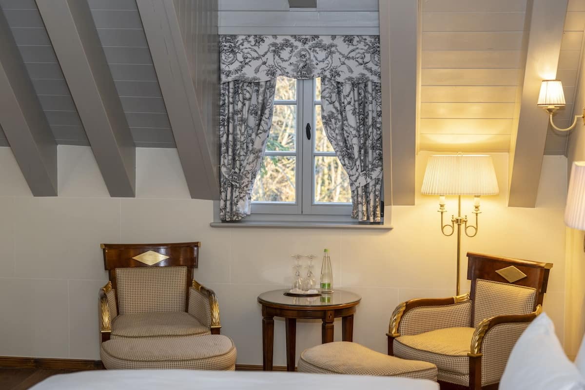 A hotel room with a bed, two upholstered chairs, a small table with a lamp, and a window with patterned curtains.
