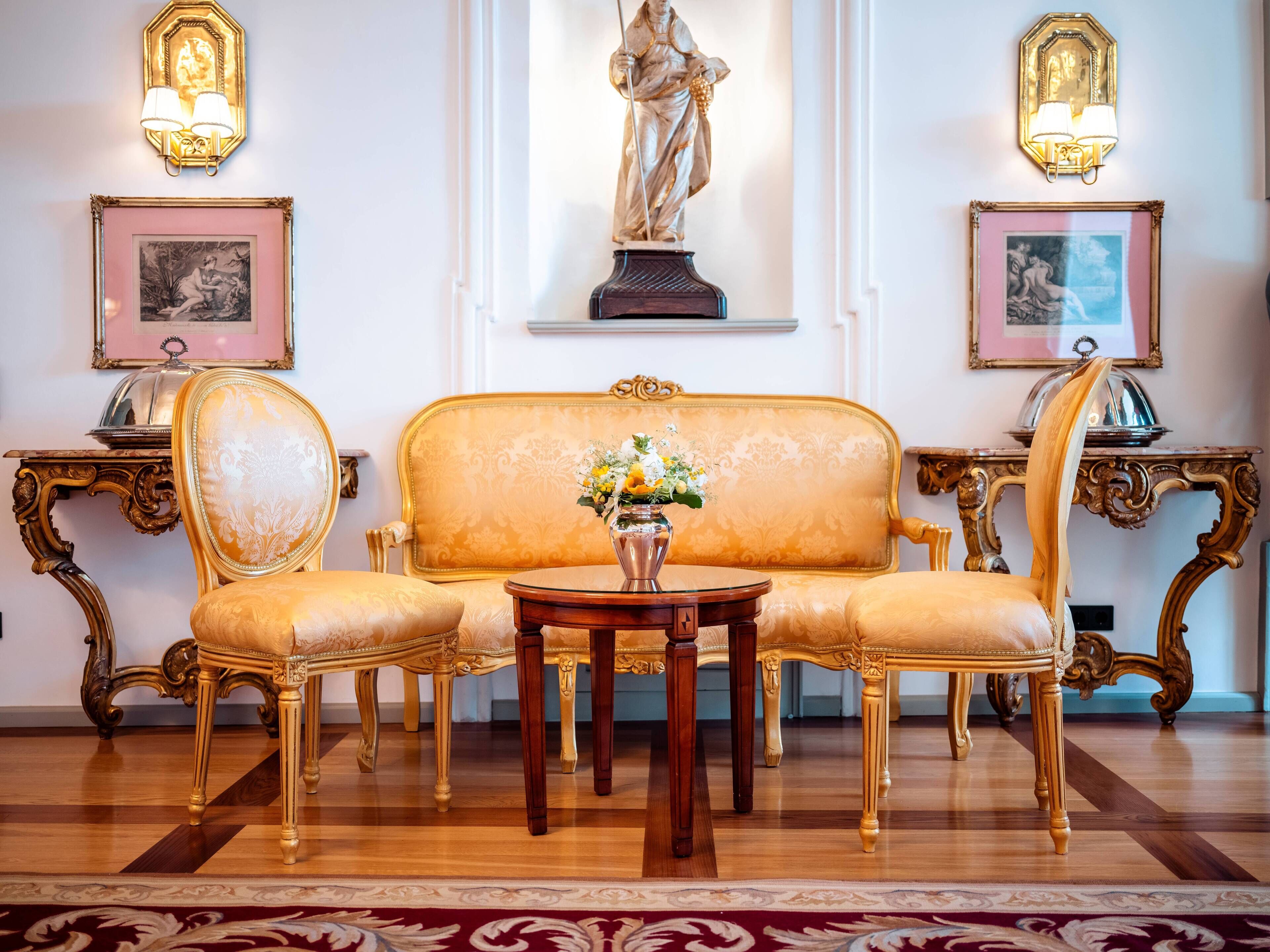 A room with a gold upholstered sofa, two matching chairs, a small wooden table with a vase of flowers, and two wall-mounted lamps.