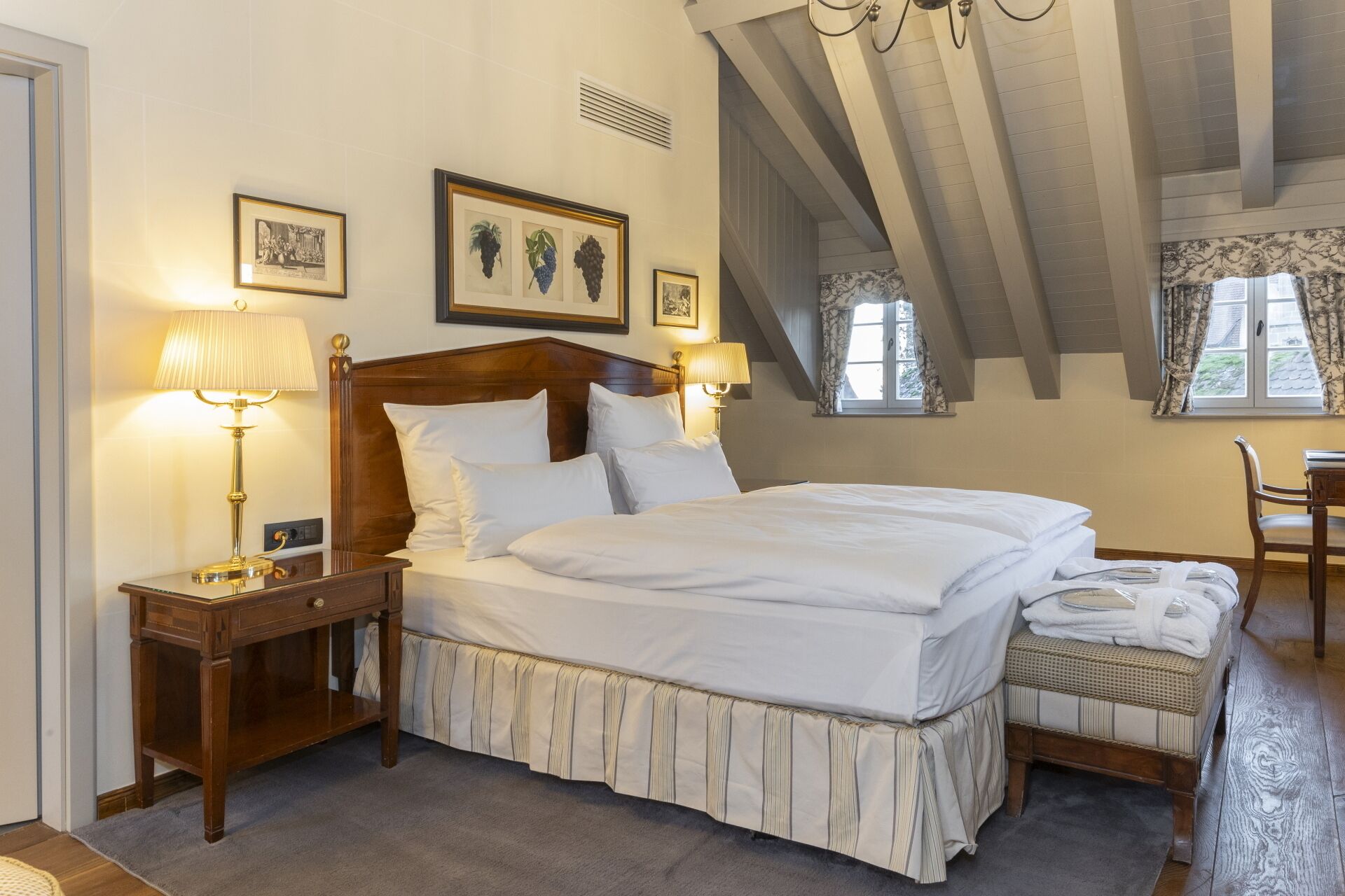 A bedroom with a large bed, two bedside tables with lamps, a wooden headboard, a window with curtains, and framed pictures on the wall.
