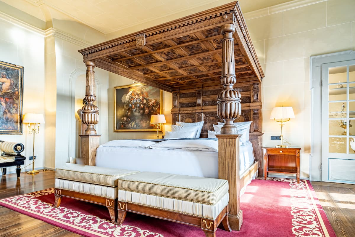 A four-poster bed with a red and white patterned rug, a bench, and a painting above the bed.