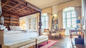 A spacious bedroom with a large bed, a sitting area with chairs, a desk, and a view of the outdoors through a window.