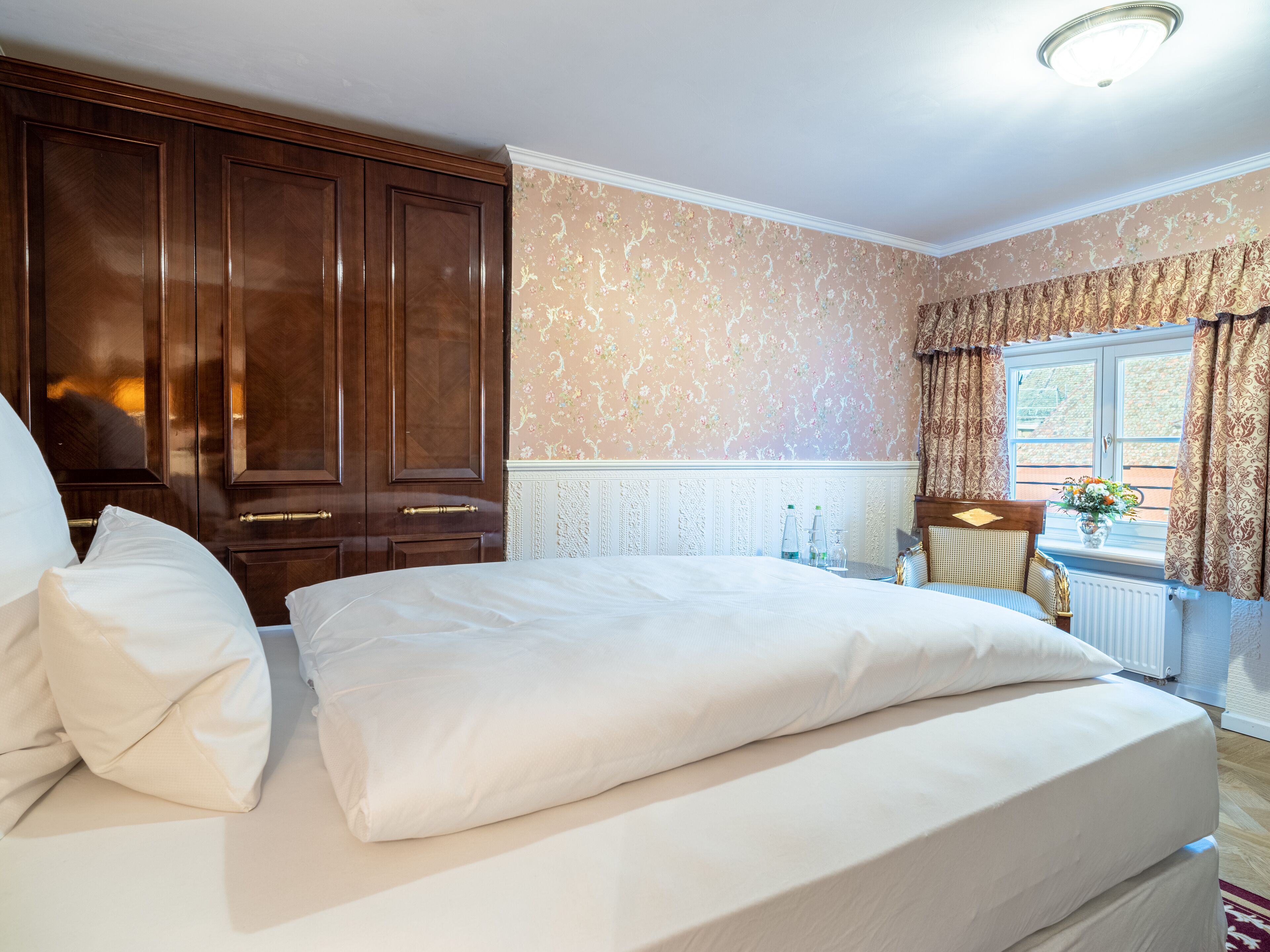 A bedroom with a large bed, wooden wardrobe, floral wallpaper, and a window with curtains.
