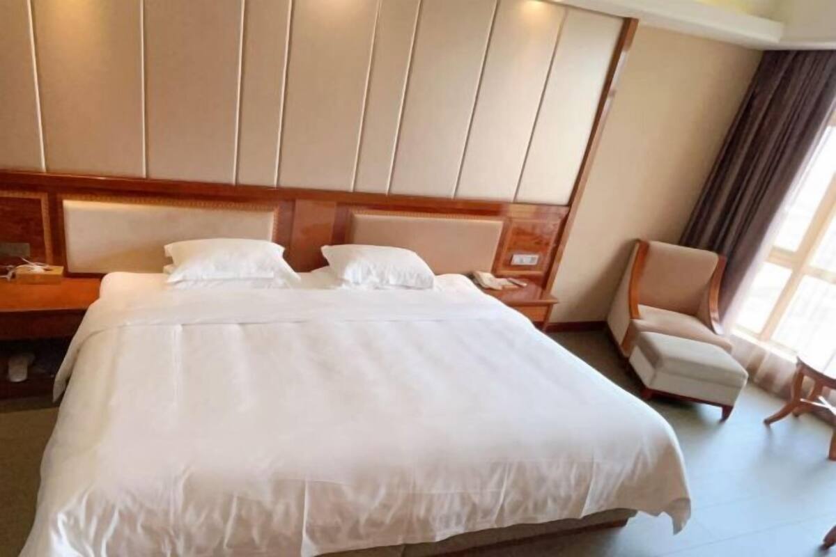 Une chambre d’hôtel avec un grand lit, une tête de lit en bois, une table de chevet, une chaise et une fenêtre avec des rideaux.