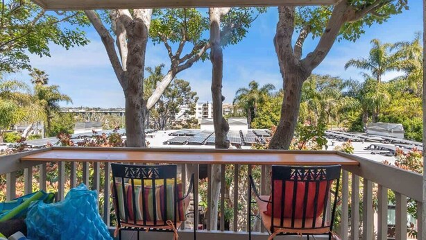 Un balcon avec deux chaises et un coussin bleu, donnant sur un parking bordé d’arbres et entouré de bâtiments.