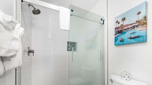 Une salle de bain moderne avec une douche à parois de verre, des toilettes avec un rouleau de papier toilette et un tableau représentant une scène de piscine accroché au mur.