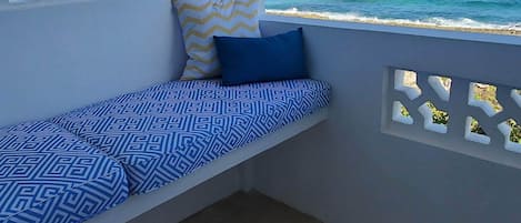 A balcony with a blue patterned sofa, a yellow and blue pillow, and a view of the ocean.