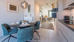 A modern kitchen with a dining area, featuring a round table with chairs, a pendant light, and framed artwork on the walls. - Smeysea - stylish apartment in Sint-Idesbald (Koksijde)