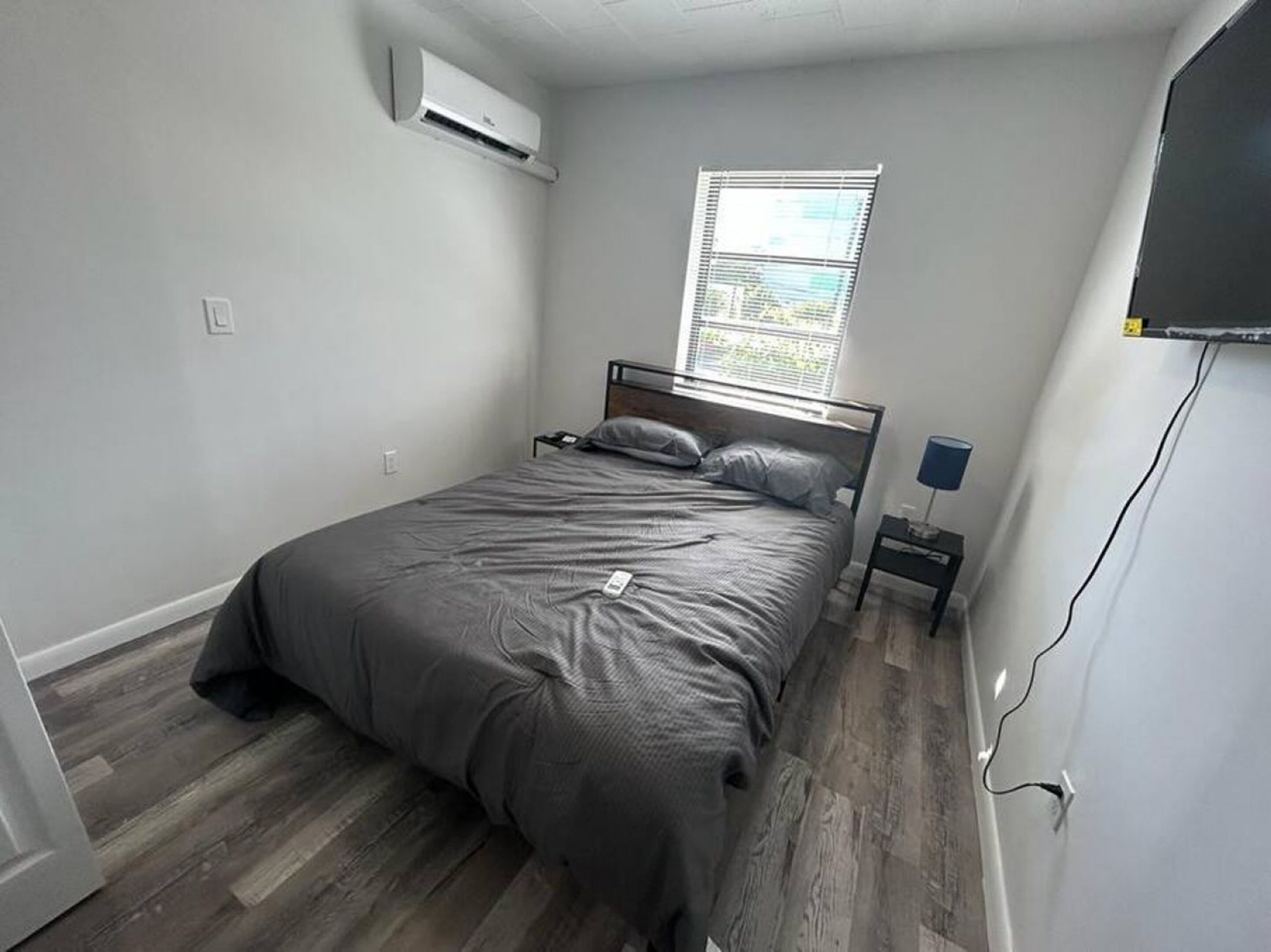 A bedroom with a bed, a window, an air conditioner, a nightstand, and a wall-mounted TV.