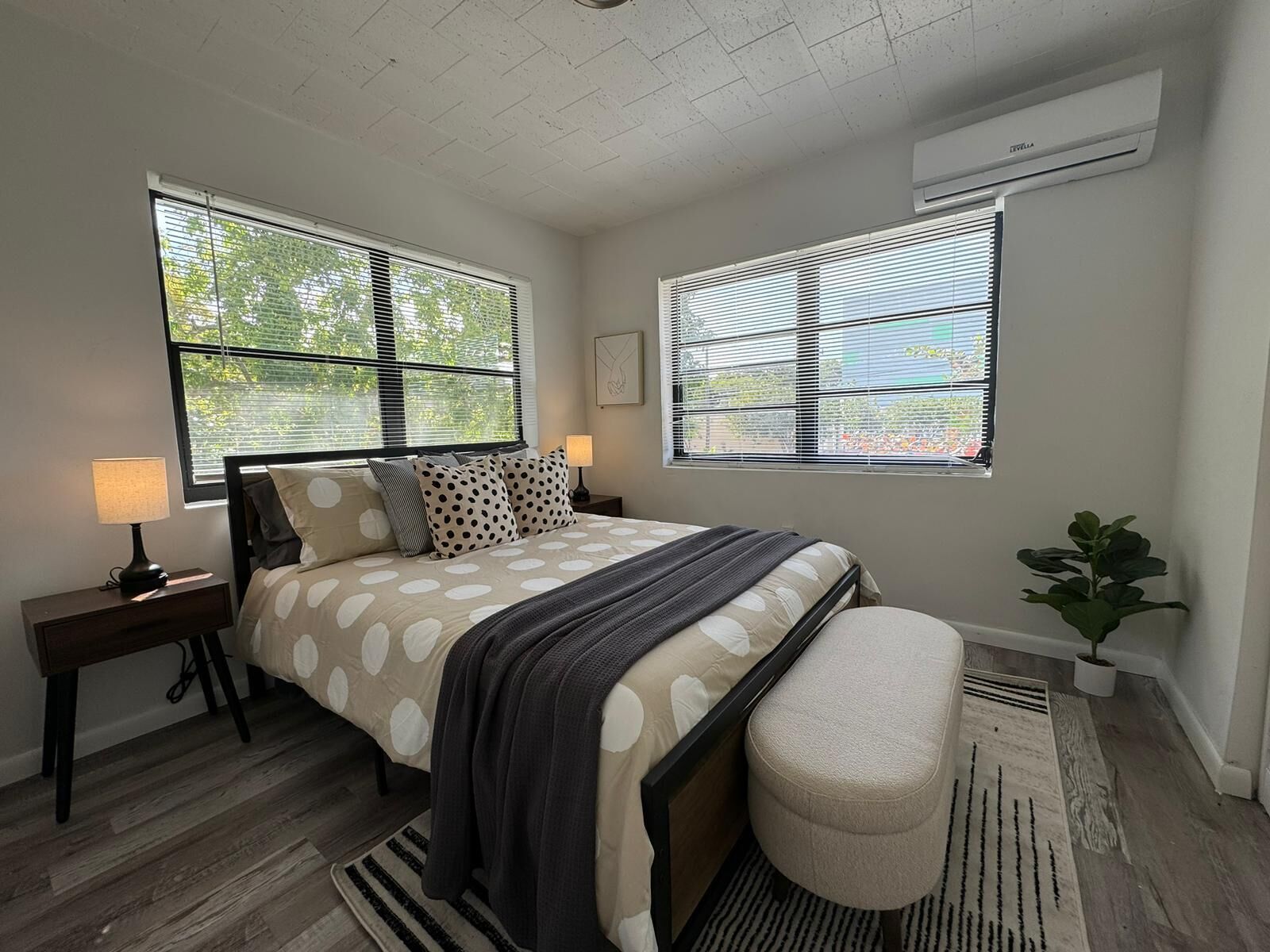 A bedroom with a bed, two bedside tables, a nightstand, a potted plant, and a window with blinds.