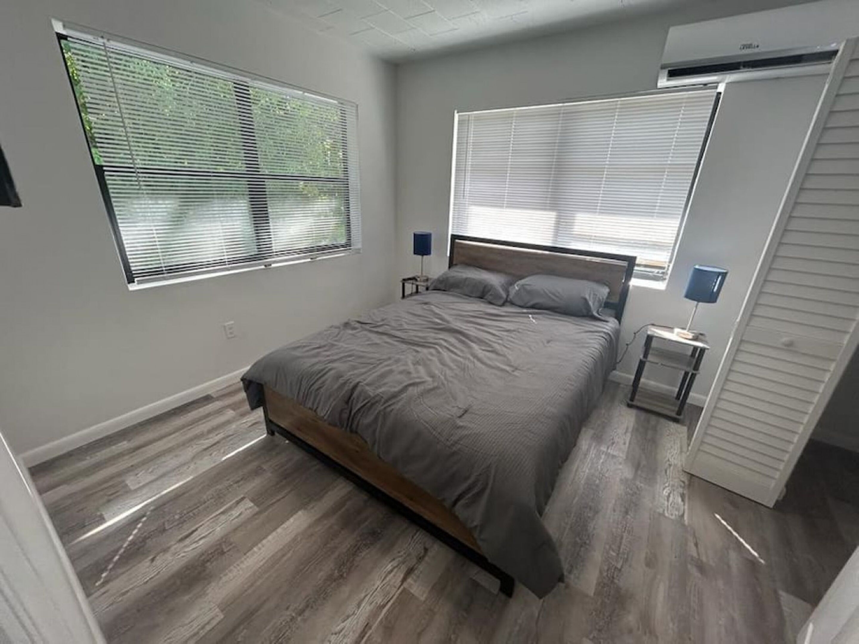 Ein Schlafzimmer mit einem Bett, zwei Nachttischen, einem Kleiderschrank und zwei Fenstern mit Jalousien.