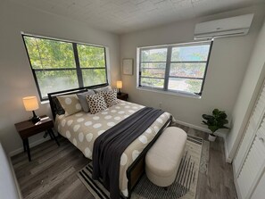 A bedroom with a bed, two bedside tables, a nightstand, a bench, a potted plant, and two windows with blinds. - Building next to CocoWalk & Brickell (Miami)