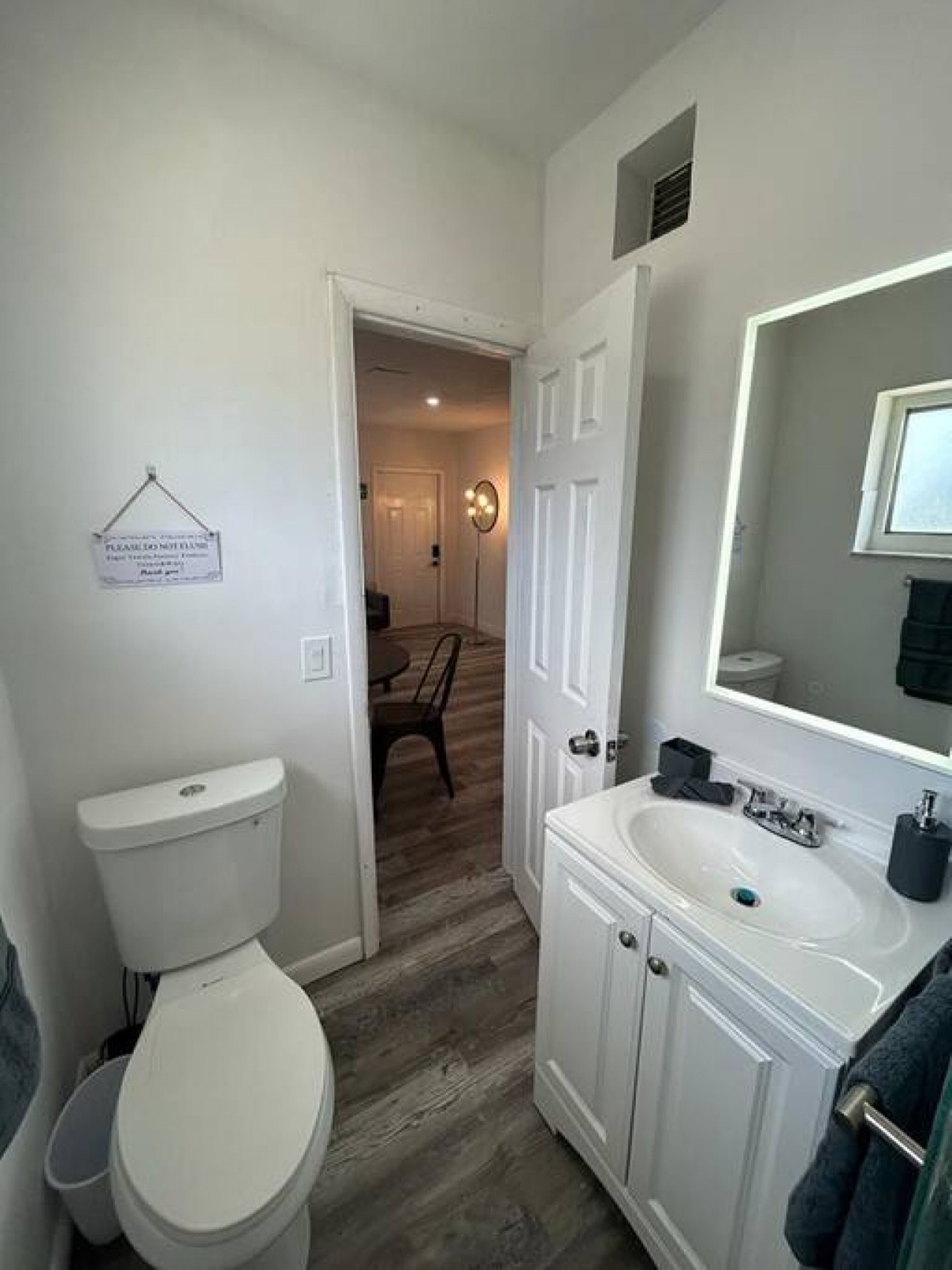 Une salle de bain avec des toilettes, un lavabo et un miroir. Il y a un panneau sur le mur et une porte qui donne sur une autre pièce.