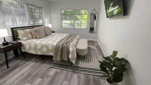 A bedroom with a bed, bedside table, lamp, windows, and a potted plant.