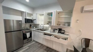 A compact kitchen with a stainless steel refrigerator, microwave, and stove. It features white cabinetry, a wooden floor, and a small dining area with a table and chairs.