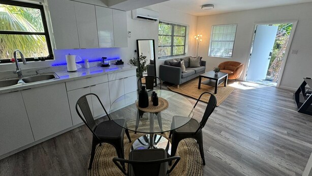 A modern kitchen with a dining area, a living room with a sofa and armchairs, and a large window with blinds.