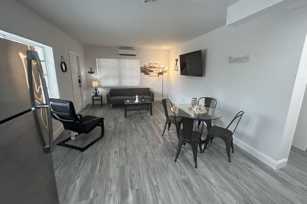 A modern living room with a sofa, coffee table, and dining table with chairs. There is a TV mounted on the wall and a ceiling light fixture.