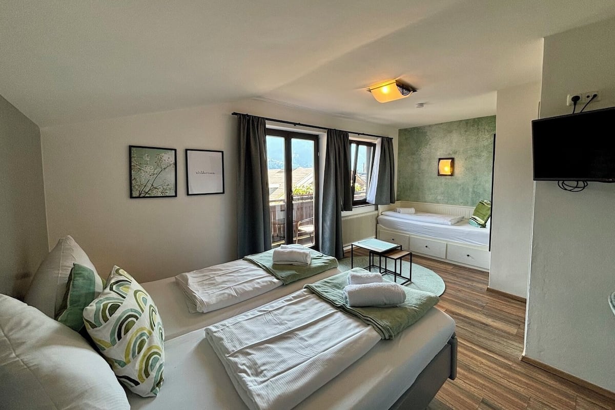 Quarto de hotel com cama, uma pequena mesa, um sofá, uma janela com cortinas, televisão na parede e banheiro com banheira.