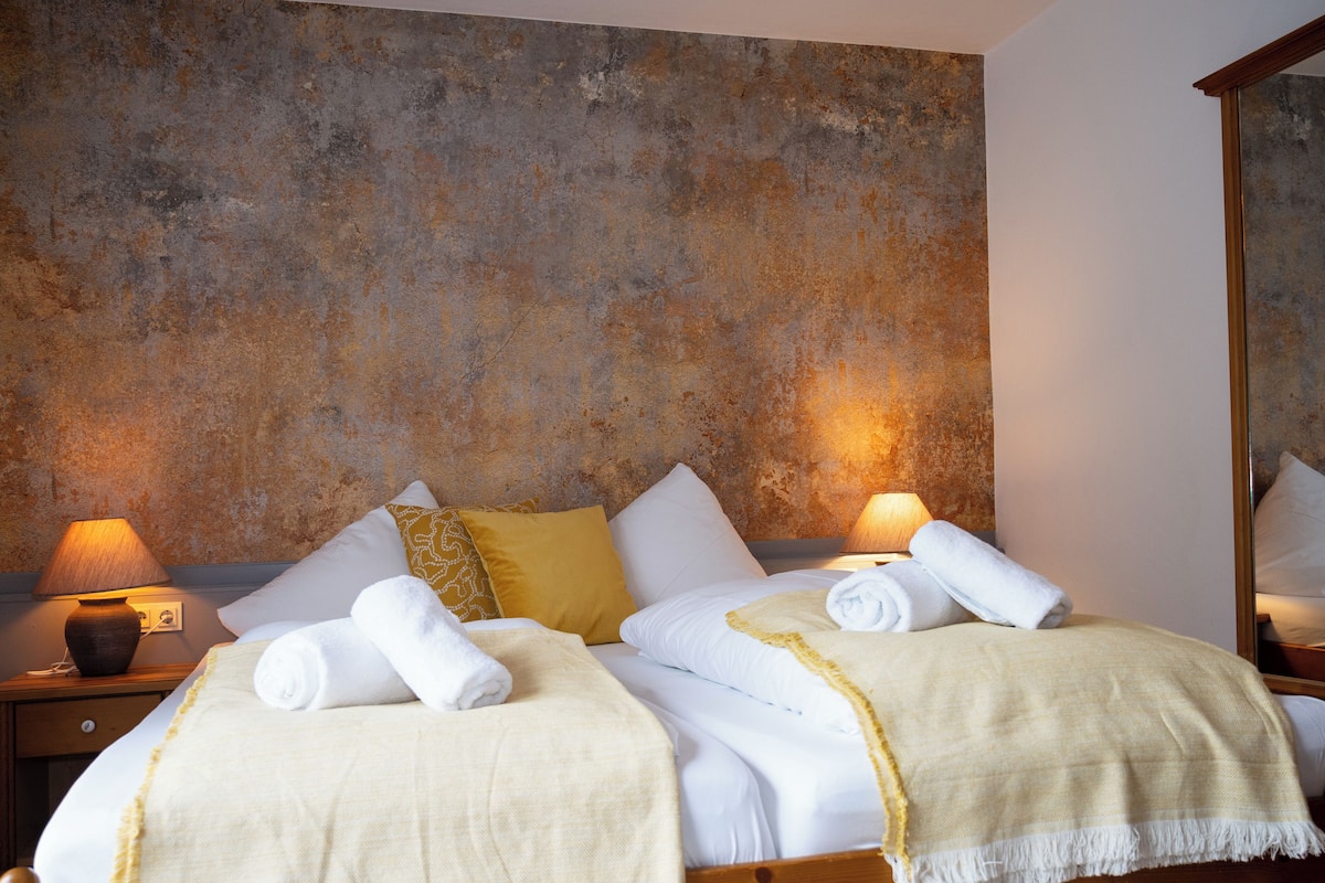 Cama bem arrumada com lençóis brancos e manta amarela, duas luminárias laterais e uma parede texturizada atrás da cama.