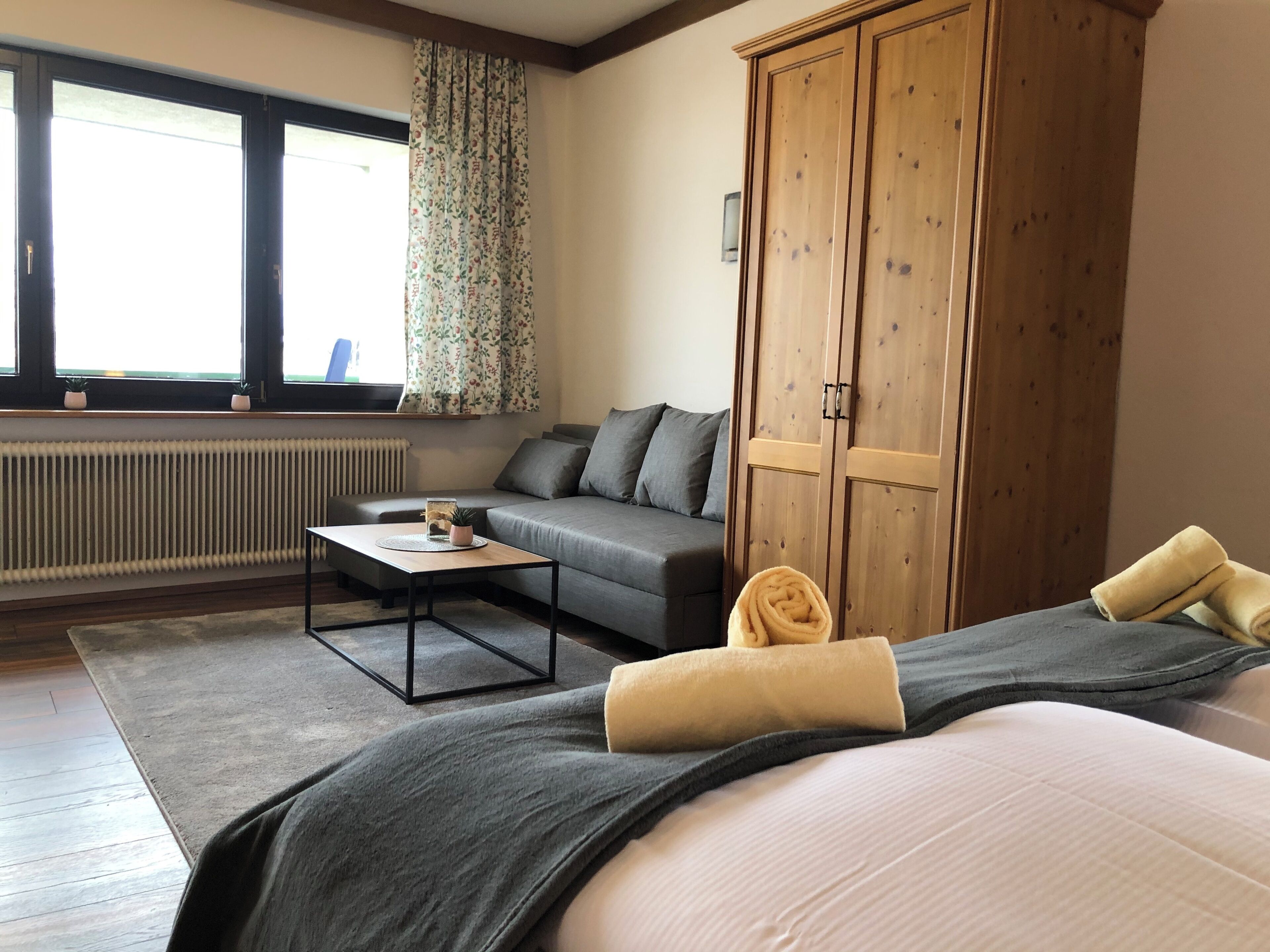 Une chambre d’hôtel avec un lit, un canapé, une armoire en bois, un radiateur et une fenêtre avec des rideaux.