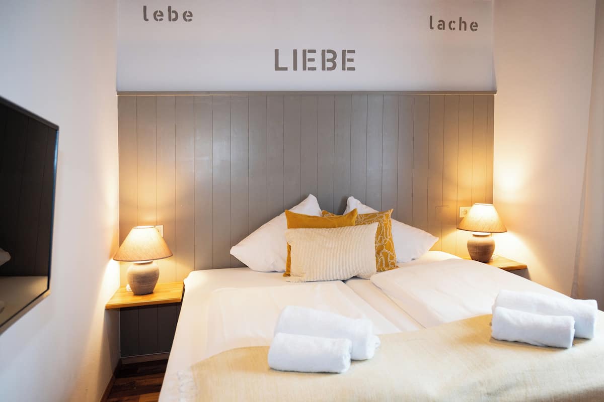Uma cama bem arrumada com lençóis brancos e duas almofadas, um abajur e uma parede com a palavra 'LIEBE' ao centro.