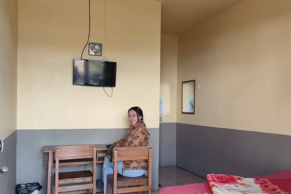 A room with a bed, a wooden chair, a table, a television mounted on the wall, and a framed picture on the wall.