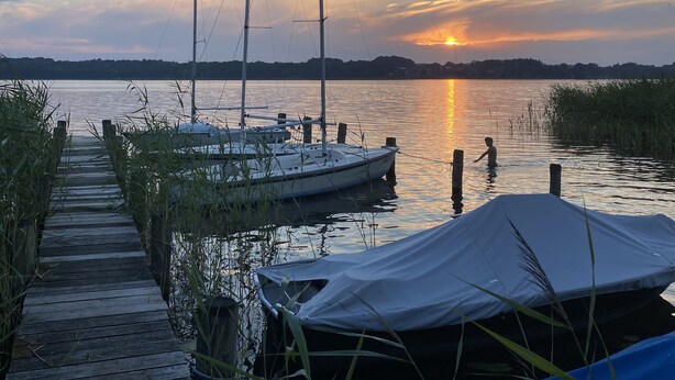 A wooden dock leading to a calm body of water with boats docked and a sunset in the background. - Apartment flying stag (Bäk)