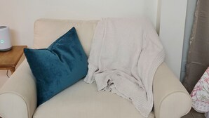 Ein Wohnzimmer mit einem beige farbenen Sofa, einem blauen Kissen, einer weißen Decke, einem Landschaftsbild und einem kleinen Tisch mit einem Gerät.