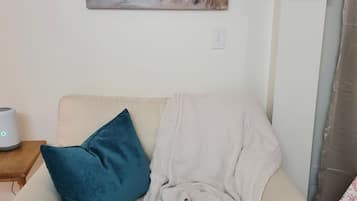 Un salon avec un canapé beige, un coussin bleu, une couverture blanche, un tableau représentant un paysage et une petite table sur laquelle se trouve un appareil.