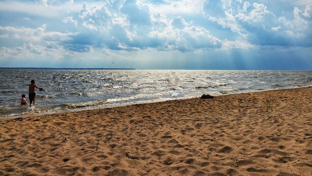 Ein Strand mit Sand und Wasser, bewölkter Himmel und eine Person im Wasser.