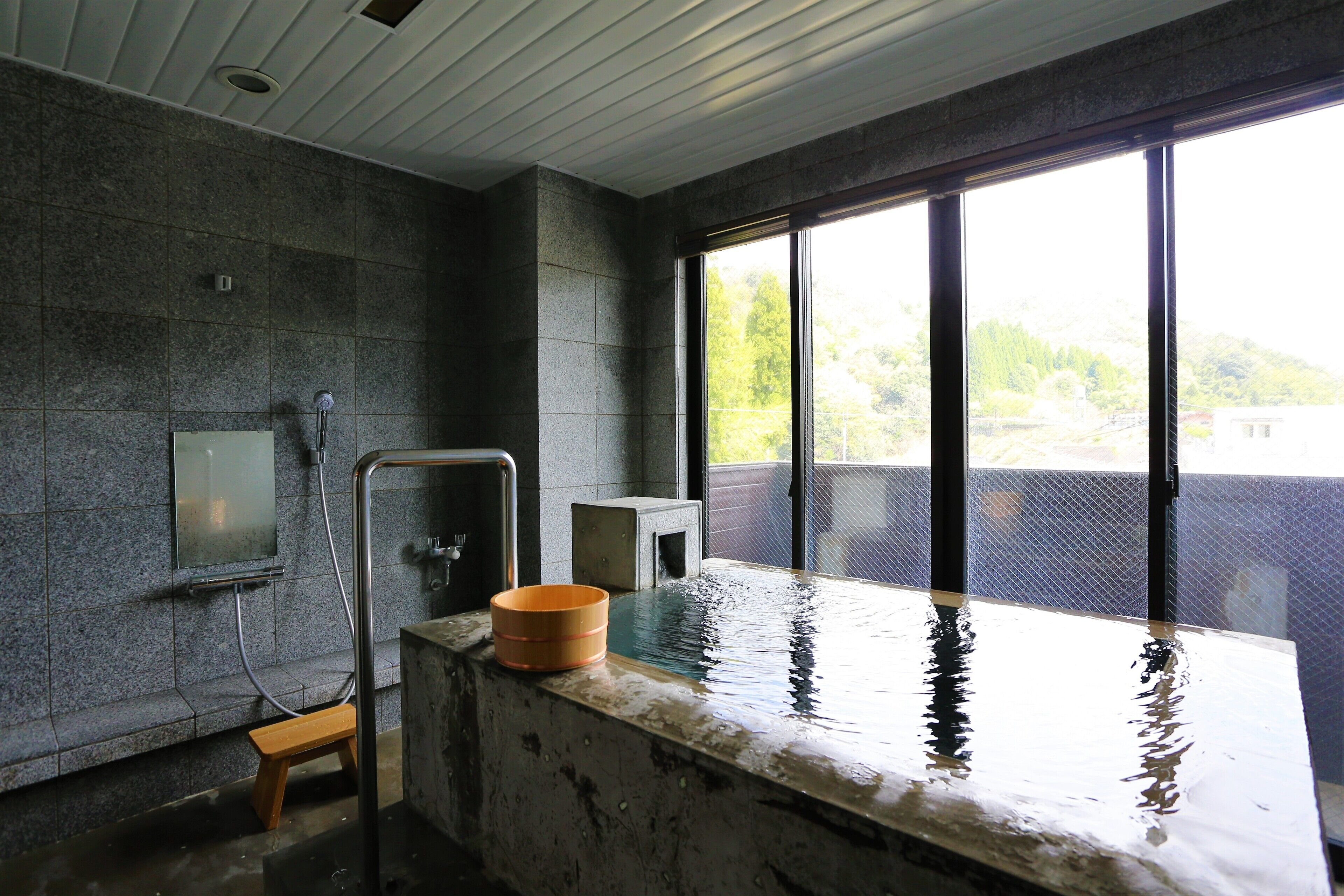 A modern bathroom with a stone wall, a wooden bench, a bathtub, and a window with a view of greenery.