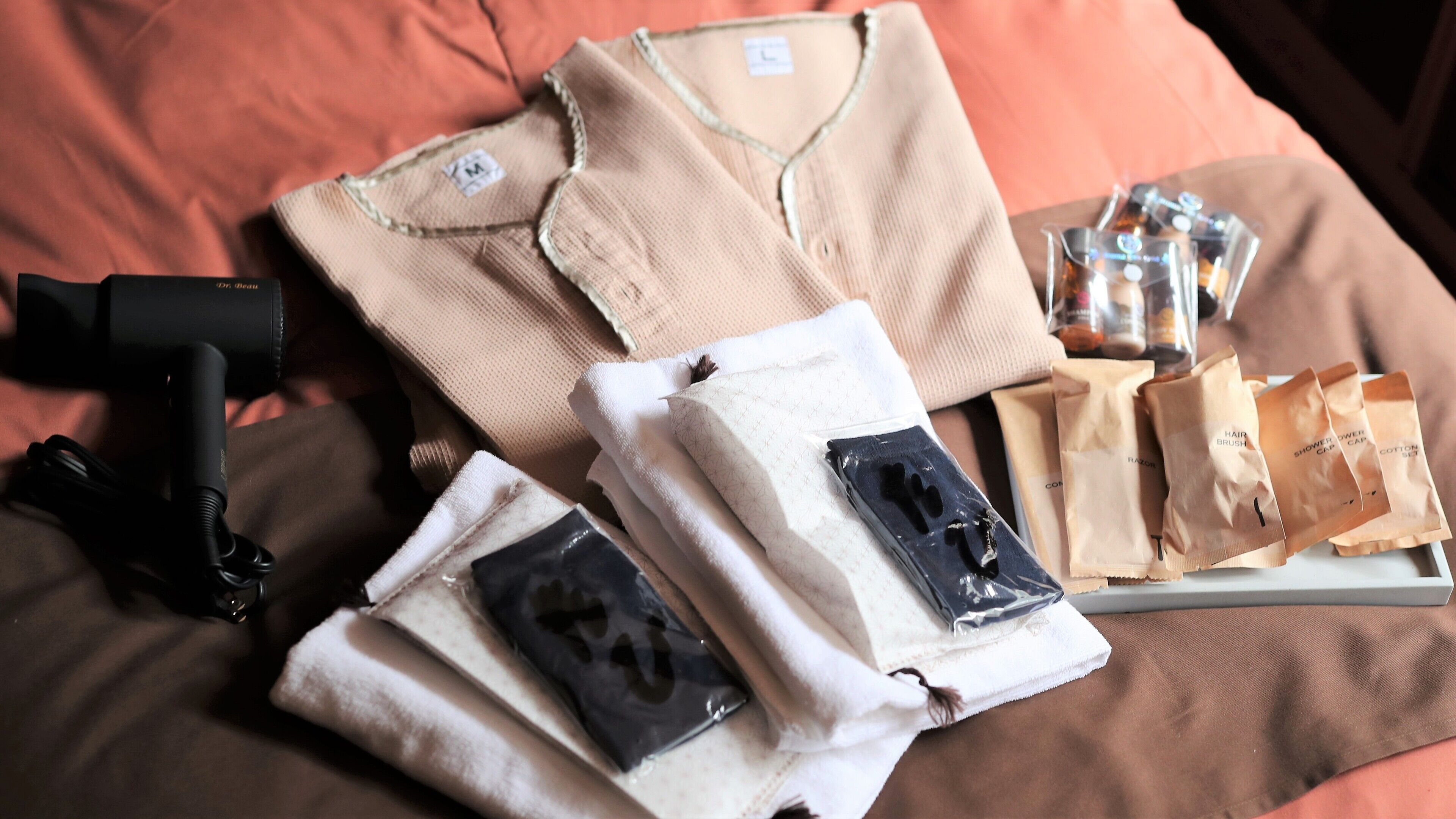 A neatly arranged bed with a brown pillow, a folded beige shirt, a black hairdryer, and several toiletry bags.