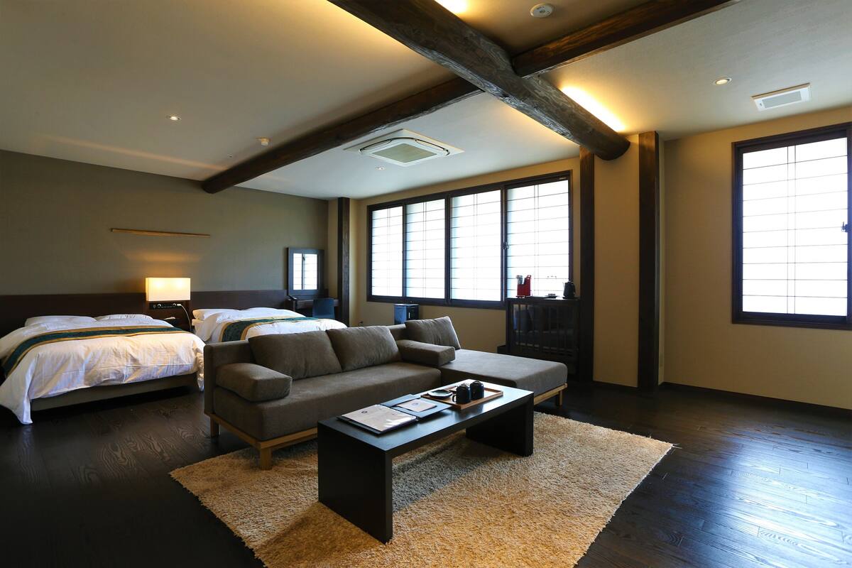 A modern hotel room with a large bed, a sofa, a coffee table, and a view of the city through large windows.