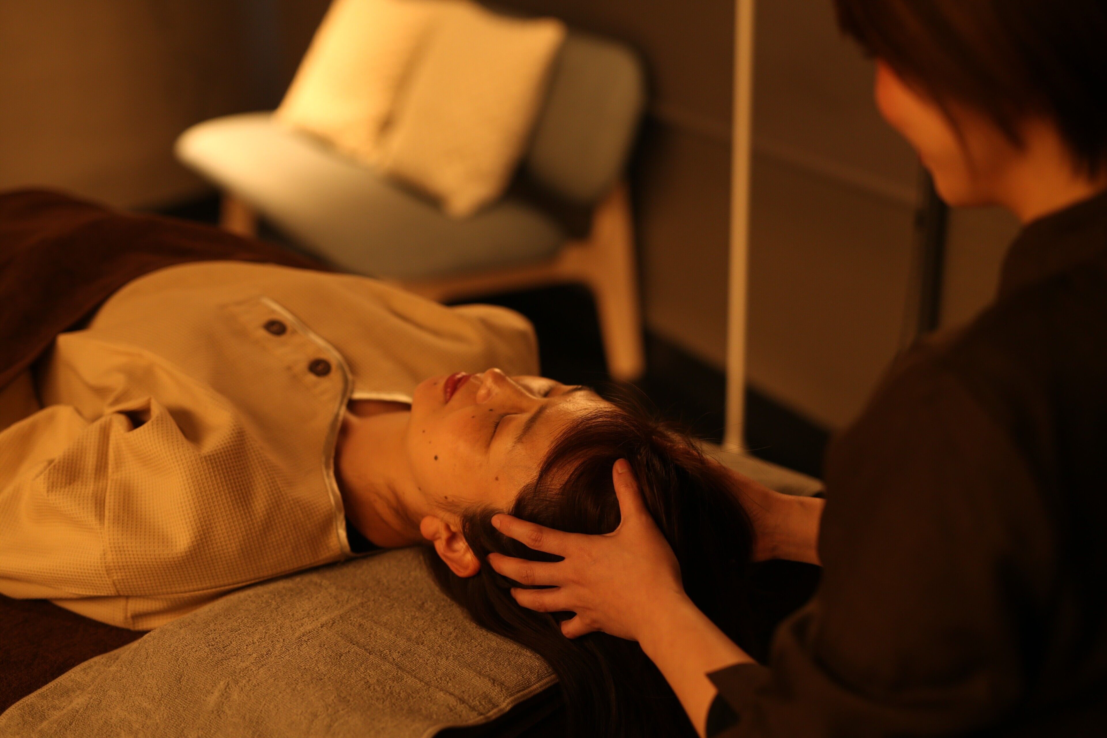 A person lying on a bed with a pillow, another person standing beside them, and a chair in the background.