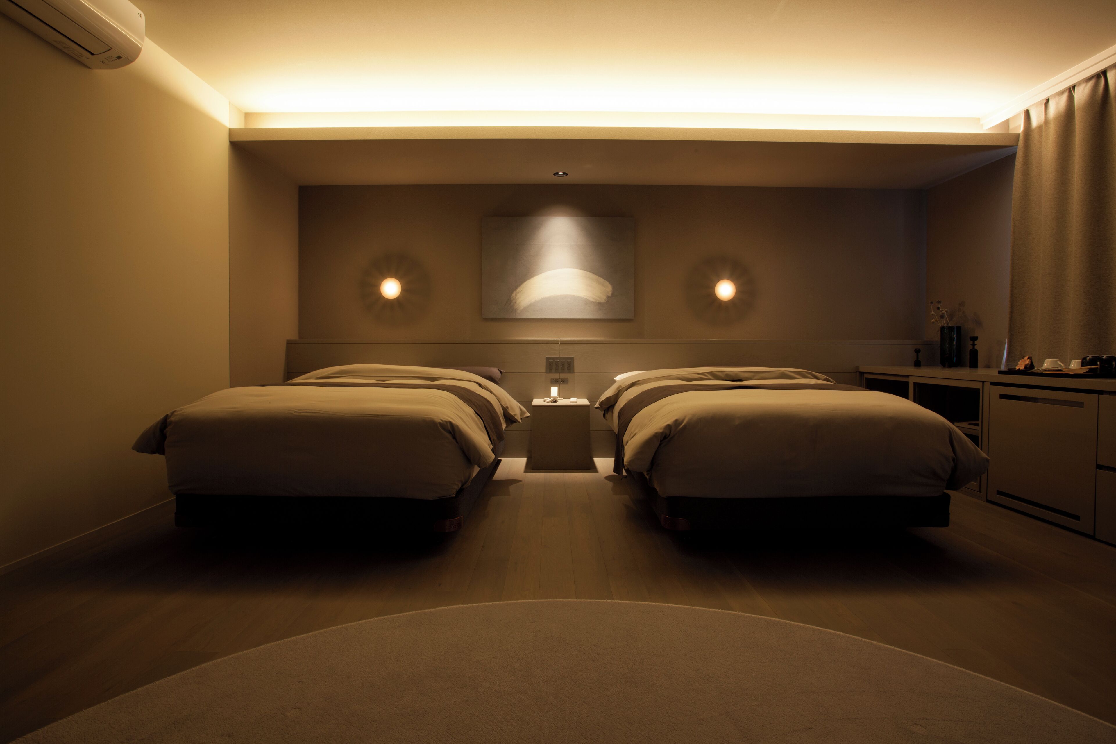 A hotel room with two beds, a nightstand, and a desk with a lamp.