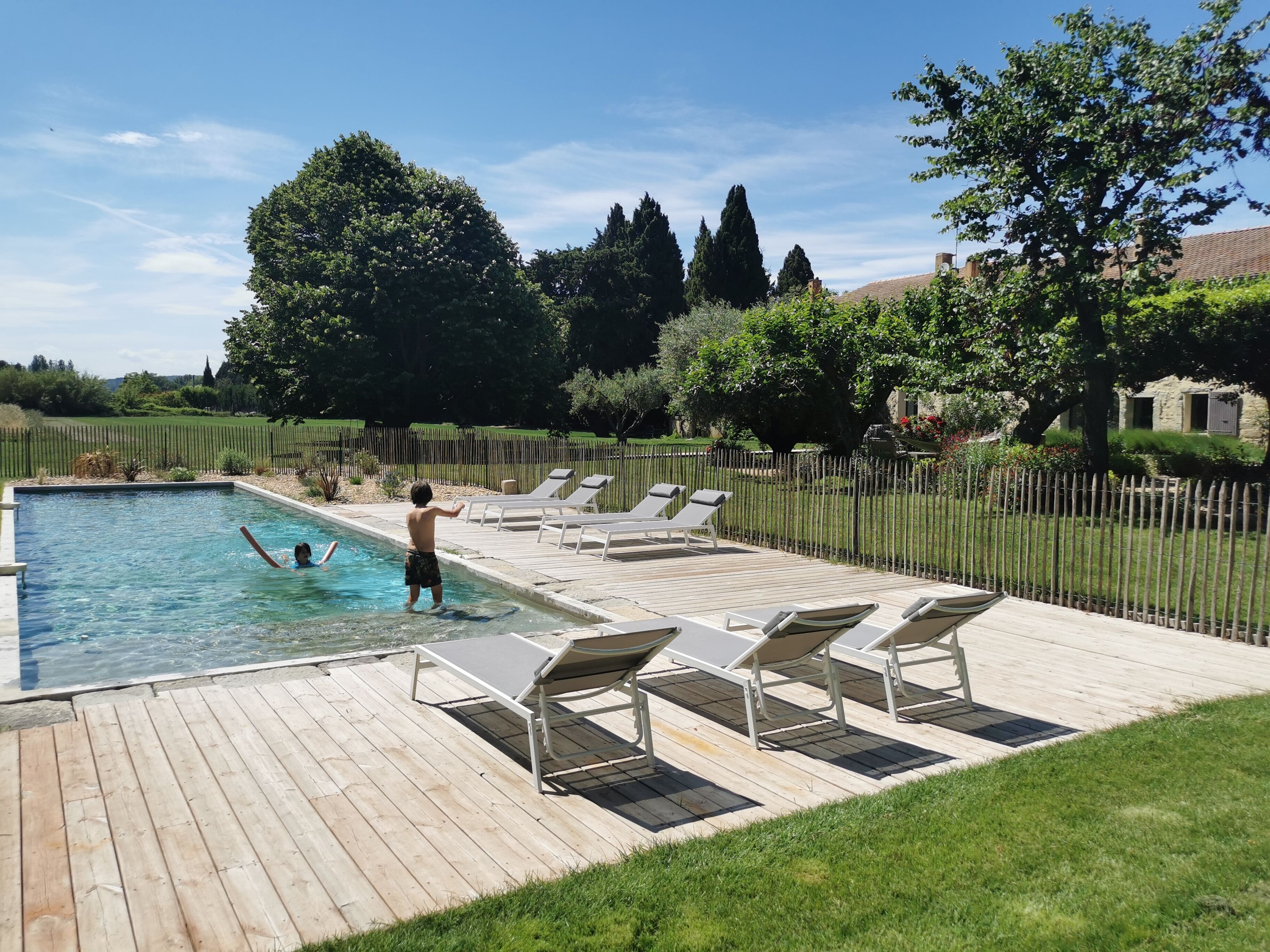 Una piscina con dos niños jugando, rodeada de sillas de descanso y una terraza de madera.