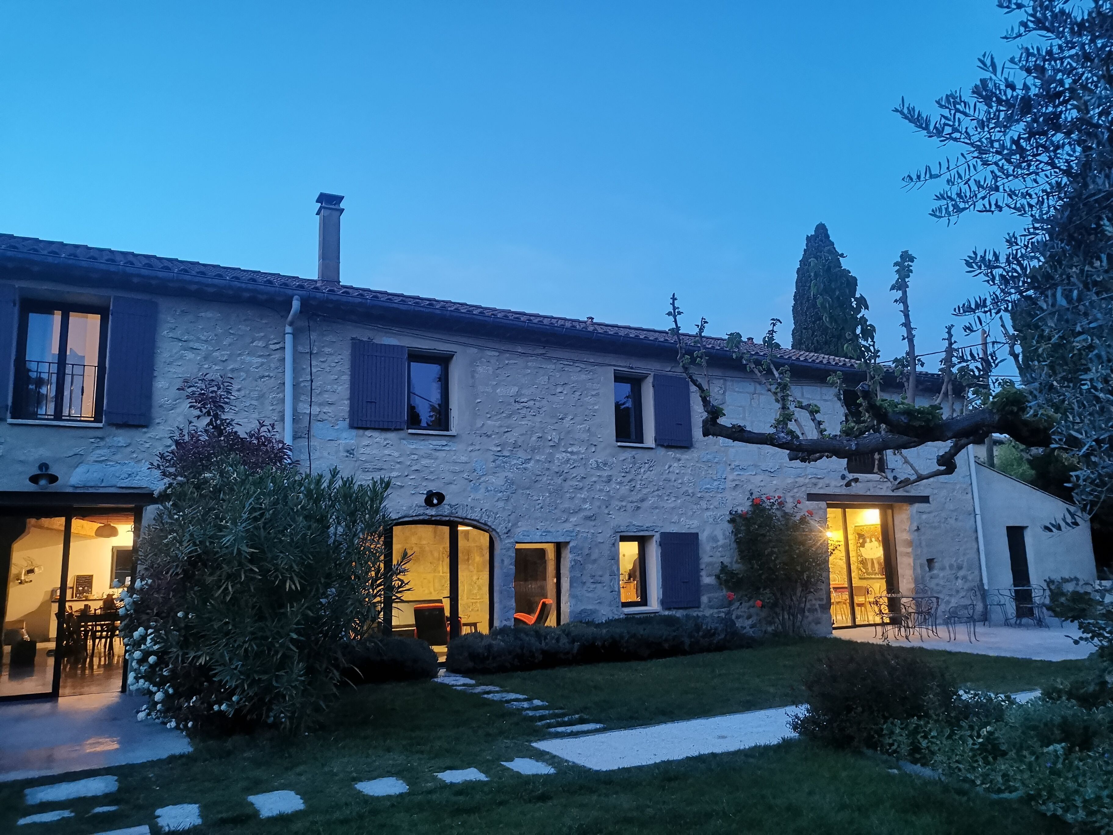 Casa de piedra con múltiples ventanas y puertas, una chimenea visible y un jardín con un sendero que lleva a la entrada.