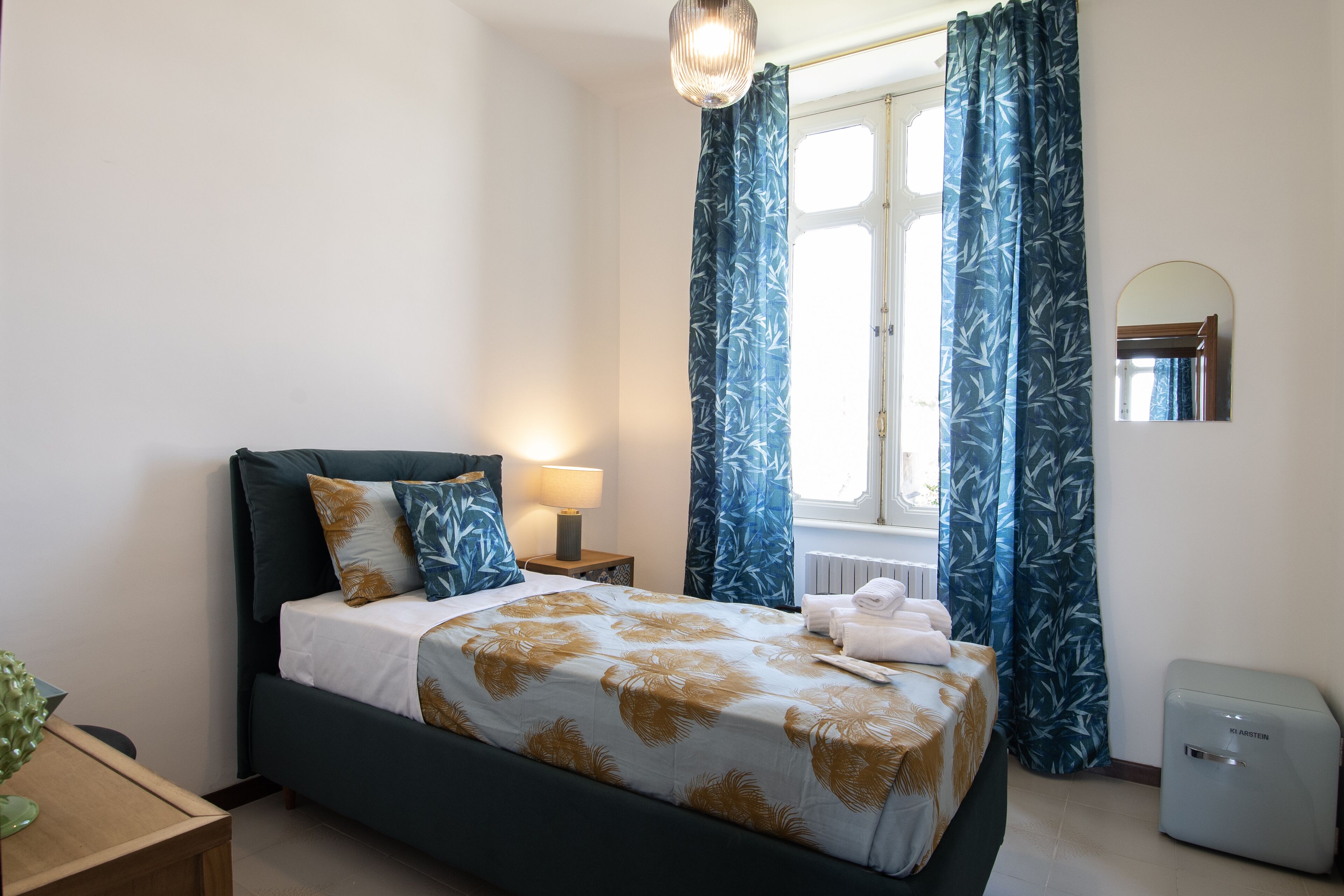 Un dormitorio ordenado con cama, mesita de noche con lámpara, radiador y pequeño calefactor.