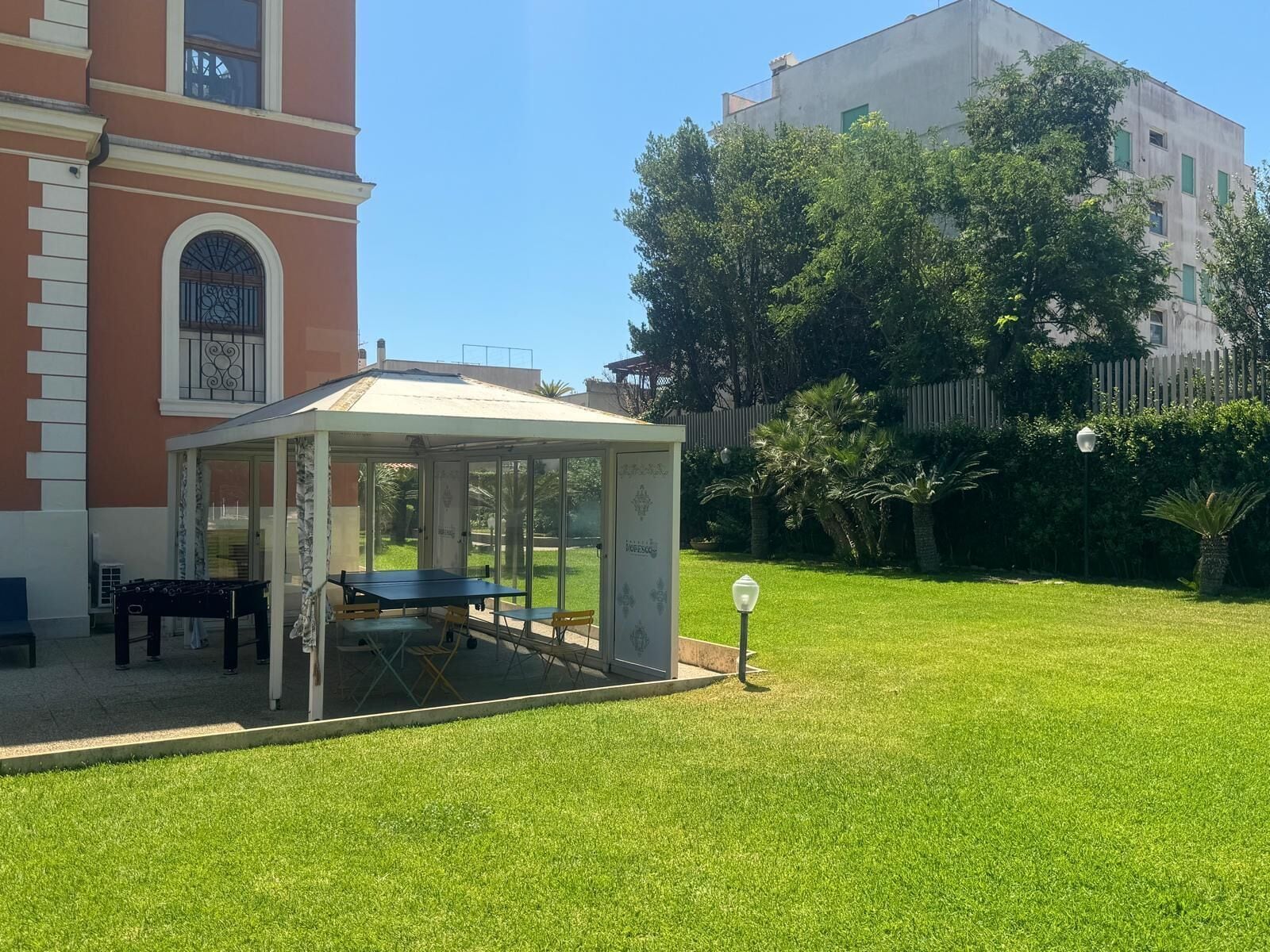 Uma área de estar coberta ao ar livre com mesa e cadeiras, um pequeno jardim e um edifício com fachada vermelha e janela com grade de ferro decorativa.