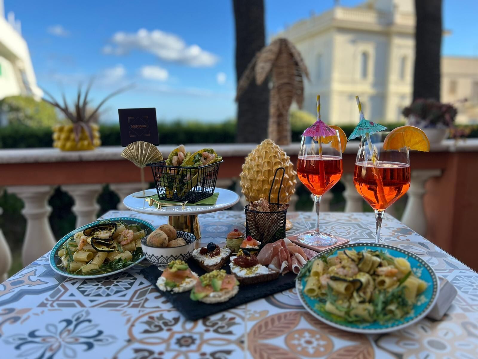 A table set with various dishes, drinks, and a menu card on a terrace with a view of a building and trees.