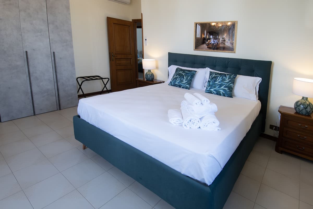 Un lit bien fait, avec du linge de lit blanc et des serviettes pliées, une tête de lit rembourrée de couleur vert foncé et un tableau encadré accroché au mur.