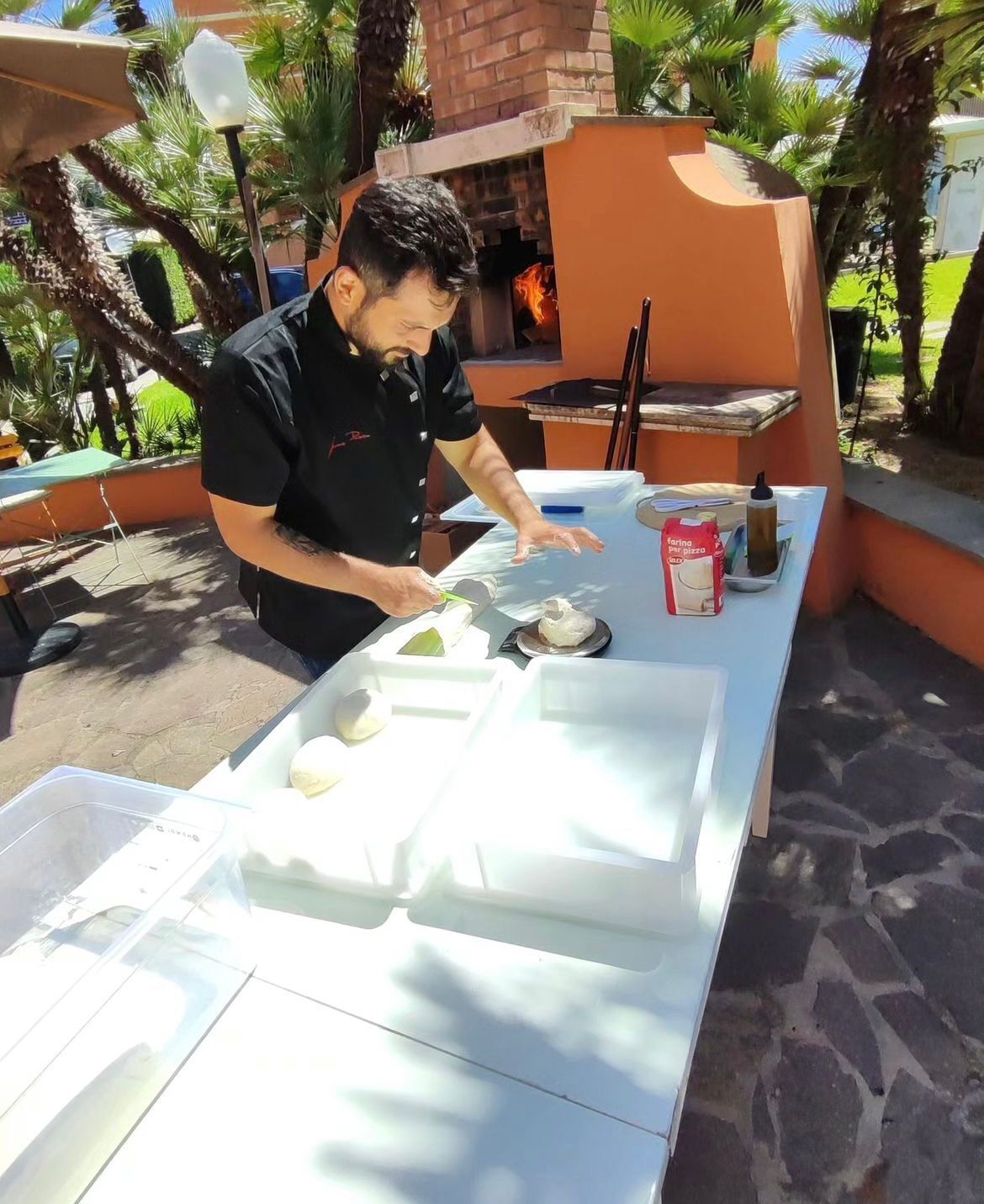Una persona preparando comida al aire libre, con una mesa, un horno de ladrillo y un césped verde al fondo.
