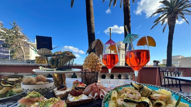 A rooftop dining setup with various dishes, including seafood, pasta, and bread, along with two glasses of red wine.