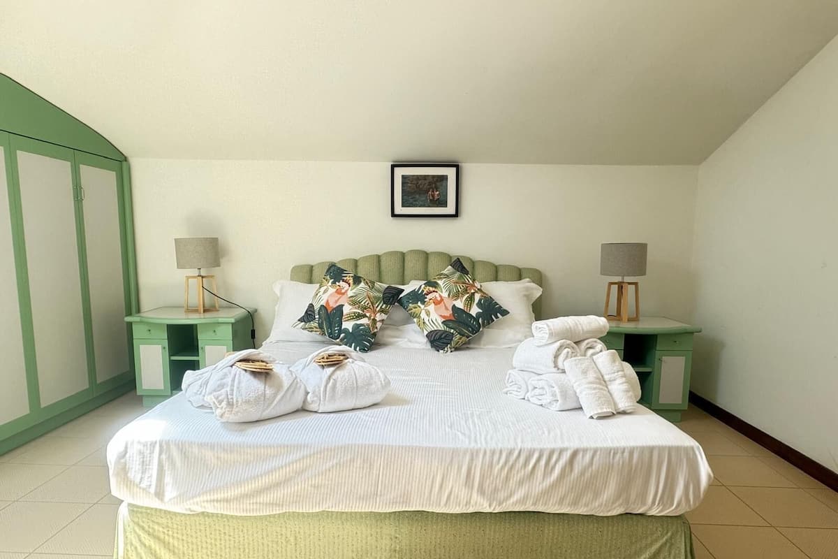 Un lit bien fait, avec du linge de lit blanc et une housse de lit verte, deux tables de chevet avec des lampes et un tableau encadré au-dessus du lit.