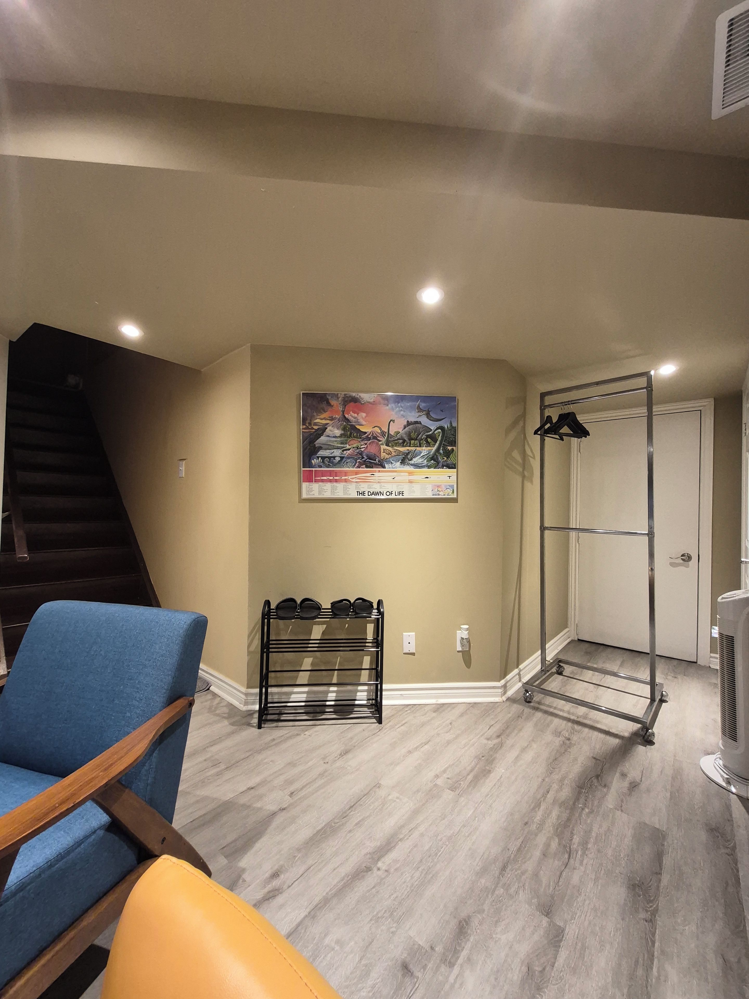 A room with a wooden staircase, a blue chair, a shoe rack, a door, and a wall-mounted picture.