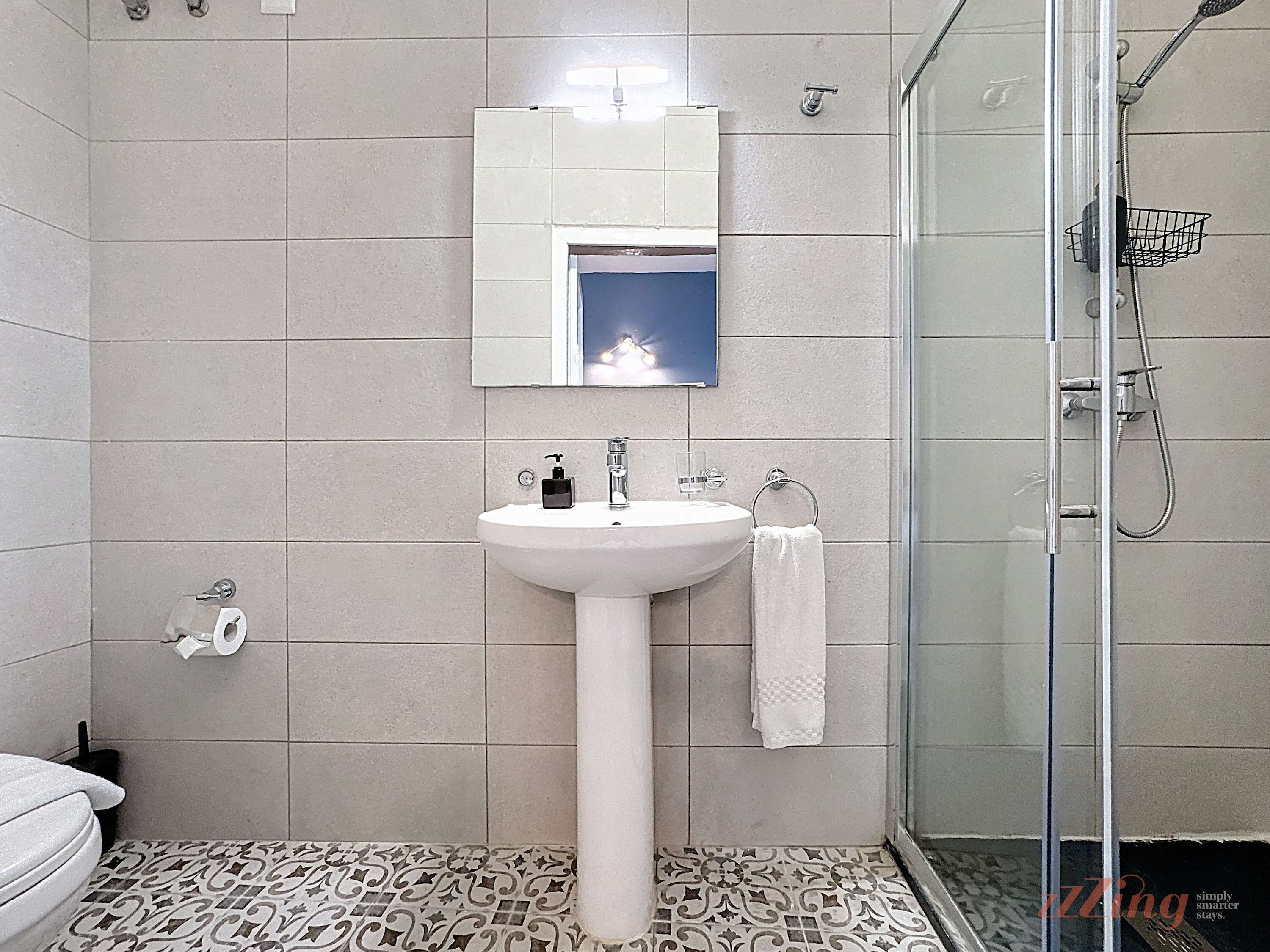 Un baño con un lavamanos blanco, un espejo, una cabina de ducha de vidrio y baldosas de suelo estampadas.