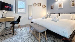 Ein Hotelzimmer mit einem Bett, einem Schreibtisch mit einem Laptop, einem Stuhl, einem Fernseher, der 'NETFLIX' anzeigt, einem Fenster und einem gemusterten Boden.
