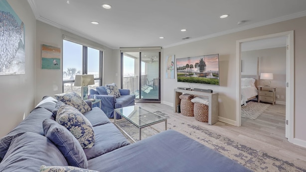 Ein modernes Wohnzimmer mit einem blauen Sofa, einem Glastisch und einem Fernseher an der Wand. Ein Balkon bietet Blick auf die Stadt.