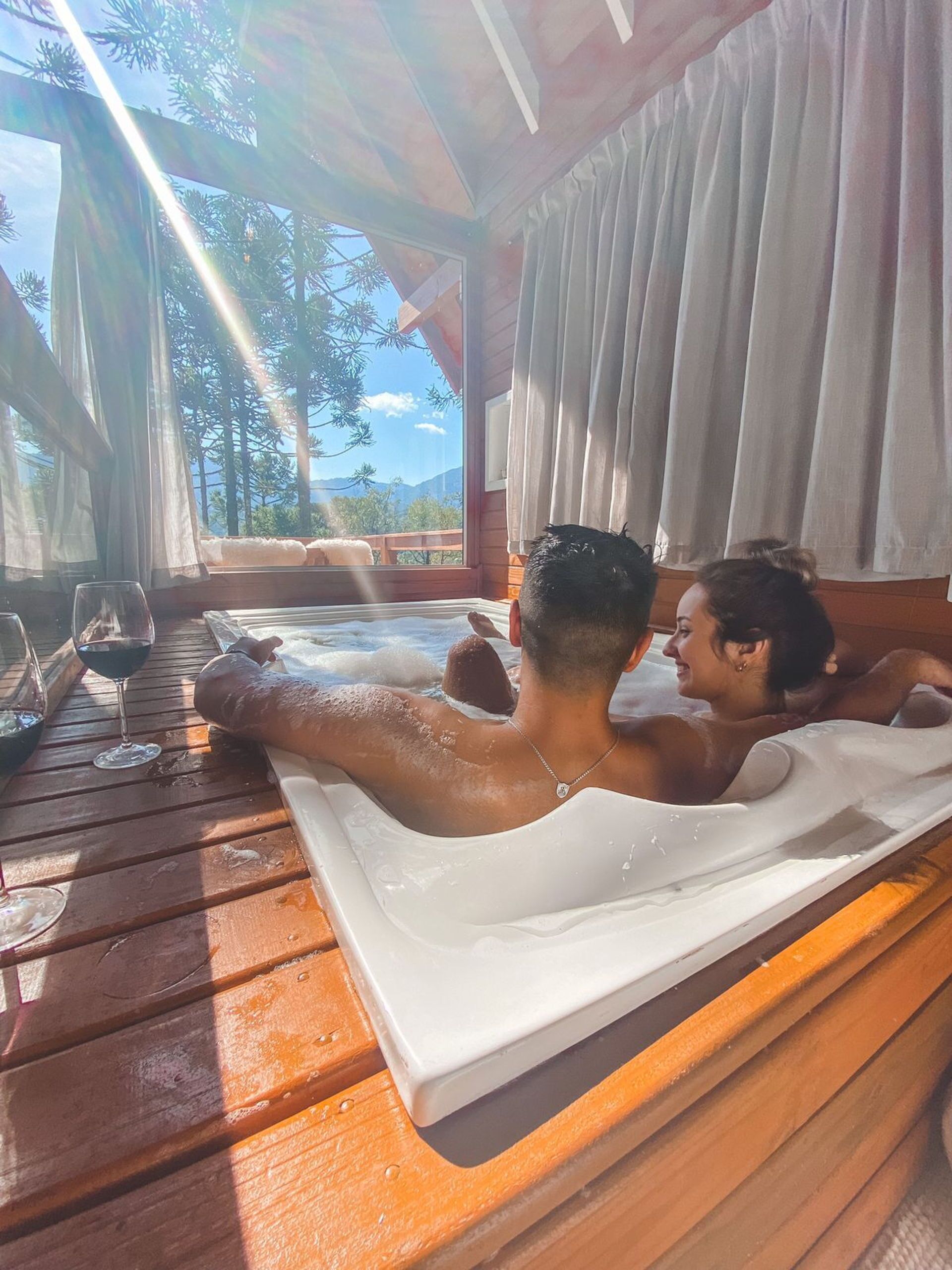 A wooden deck with a hot tub, a table with wine glasses, and a view of trees and mountains.