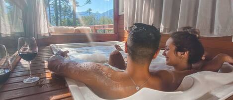 Una terraza de madera con un jacuzzi, una mesa con copas de vino y vistas a árboles y montañas.