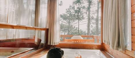 Une cabane en bois dotée d’une grande fenêtre, d’un bain à remous et offrant une vue sur les arbres.